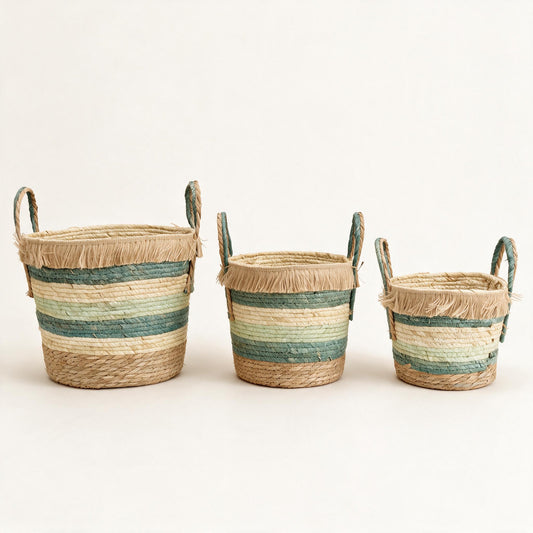Set of three coiled corn husk and seagrass nesting baskets with tan base, teal, cream and mint-sage bands, raffia fringe collar and tall looped handles — studio shot
